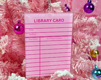 May include: Pink plastic library card ornament with the text "LIBRARY CARD" at the top and fields for "Title", "Author", "Due Date", and "Borrower's Name".
