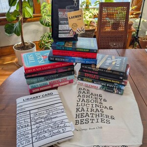 May include: A collection of books, including titles like "One in Four" and "The Nigerwife," are stacked on a wooden table. A tote bag with "The Creepy Book Club" text and a library card-themed book sleeve are also present. A black bag and a clear tag with the name "Lauren" are also visible.