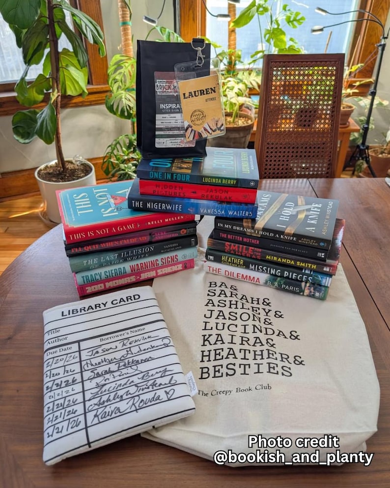 May include: A collection of books, including titles like "One in Four" and "The Nigerwife," are stacked on a wooden table. A tote bag with "The Creepy Book Club" text and a library card-themed book sleeve are also present. A black bag and a clear tag with the name "Lauren" are also visible.