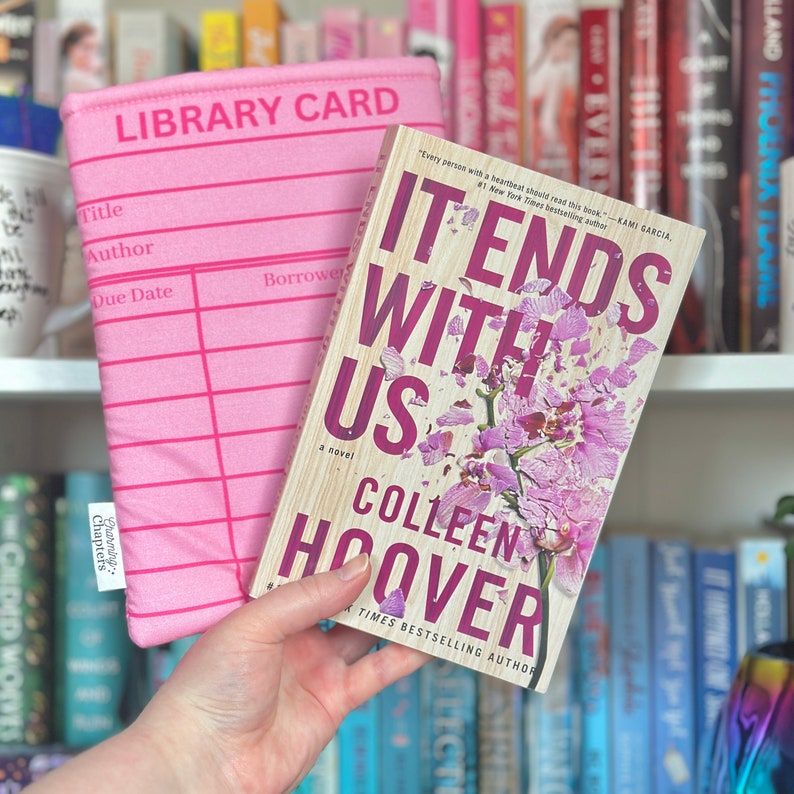 May include: A pink library card bookmark with the words "Library Card" printed on it. The bookmark is being used to hold a paperback book titled "It Ends With Us" by Colleen Hoover. The book cover features a pink orchid with petals falling off.
