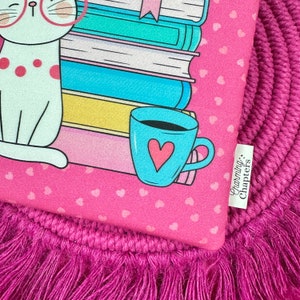 Peut inclure: Tissu rose &agrave; motifs de c&oelig;urs r&eacute;p&eacute;t&eacute;s avec un chat de dessin anim&eacute; portant des lunettes, une pile de livres et une tasse bleue avec un c&oelig;ur. Le texte "Charming Chapters" est sur une &eacute;tiquette blanche. Le tissu est sur un tapis rose &agrave; franges.