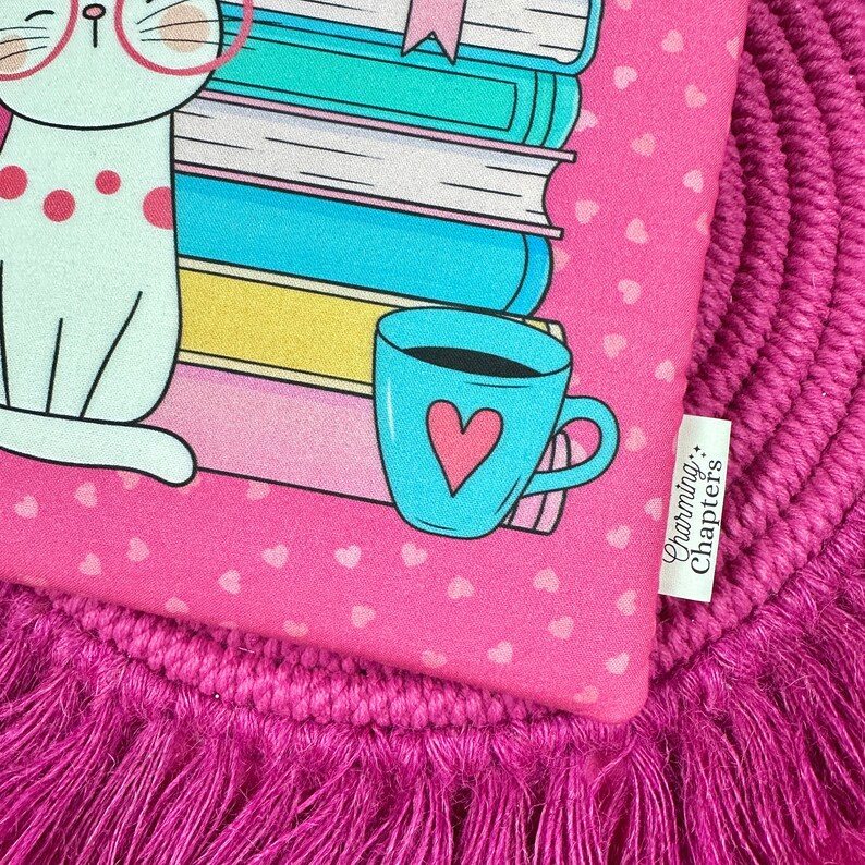 Peut inclure: Tissu rose &agrave; motifs de c&oelig;urs r&eacute;p&eacute;t&eacute;s avec un chat de dessin anim&eacute; portant des lunettes, une pile de livres et une tasse bleue avec un c&oelig;ur. Le texte "Charming Chapters" est sur une &eacute;tiquette blanche. Le tissu est sur un tapis rose &agrave; franges.
