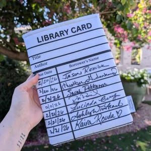 May include: A white library card-themed book sleeve with black lines and text. The text includes "LIBRARY CARD", "Title", "Author", "Due Date", and "Borrower's Name". Handwritten names and dates fill the card. The sleeve is held in front of a blurred outdoor background.