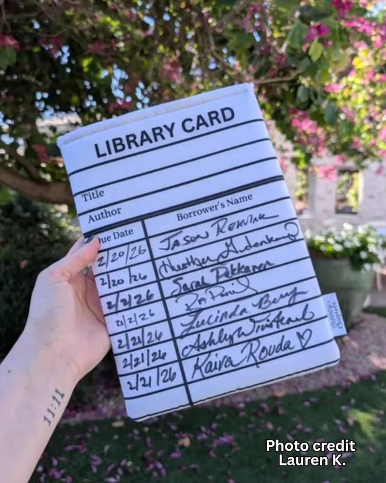 May include: A white library card-themed book sleeve with black lines and text. The text includes "LIBRARY CARD", "Title", "Author", "Due Date", and "Borrower's Name". Handwritten names and dates fill the card. The sleeve is held in front of a blurred outdoor background.