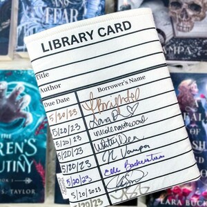 Foam padded book cover laying flat on top of a variety of books to make a dark background.  The book sleeve has a library card design and can be used at bookish events. This photo shows the book sleeve filled with dates and signatures from authors.