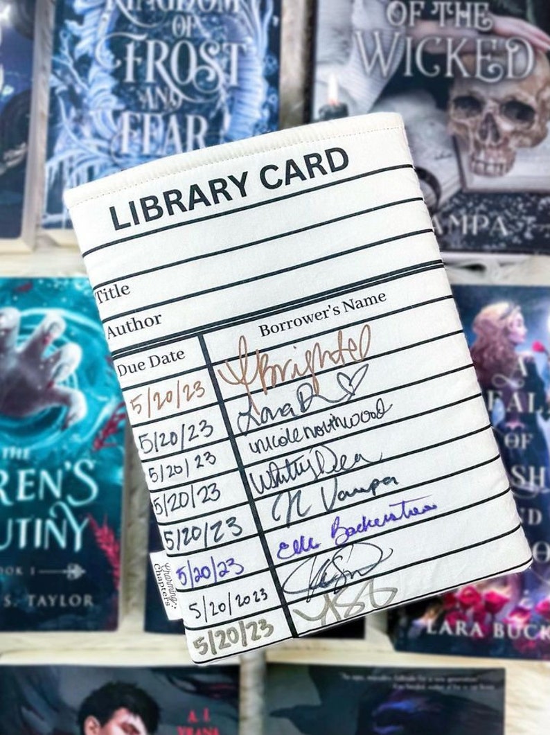 Foam padded book cover laying flat on top of a variety of books to make a dark background.  The book sleeve has a library card design and can be used at bookish events. This photo shows the book sleeve filled with dates and signatures from authors.