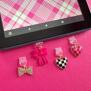 May include: Four decorative charms with gold-coloured clips. The charms include a rhinestone bow, a pink velvet bow, a black and white chequered heart, and a pink and white chequered heart. The background is a bright pink surface.