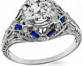 May include: A vintage-style engagement ring with a large, round, clear diamond center stone. The ring is set in platinum and features a delicate filigree design with smaller diamonds and blue sapphires.