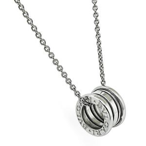 May include: A silver-toned metal necklace featuring a cylindrical pendant with a multi-layered, ribbed design. The word BVLGARI is engraved on the outer edges of the pendant, which hangs from a delicate, polished silver-toned link chain.
