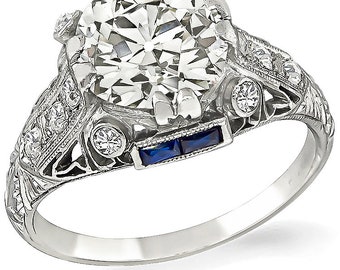 Puede incluir: Un anillo de compromiso de estilo vintage con un gran diamante redondo y claro en el centro. El anillo está engastado en platino y presenta un delicado diseño de filigrana con diamantes más pequeños y dos zafiros azules.