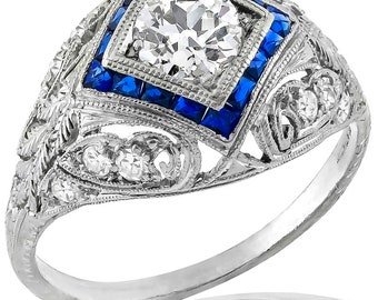 May include: A vintage engagement ring with a large, round, brilliant-cut diamond center stone surrounded by a square halo of blue sapphires. The ring has a delicate, filigree design with smaller diamonds set in the band.