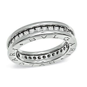May include: A silver-toned metal ring featuring a central channel set with a row of small, clear, round-cut stones. The outer edges of the band are engraved with the text BVLGARI, repeating along the circumference of the polished metal surface.