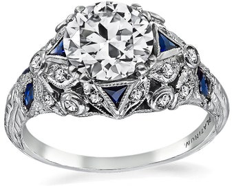 May include: A platinum engagement ring featuring a large, round, clear diamond centre stone. The ring is set with smaller diamonds and blue sapphires in a delicate, intricate design. The band is engraved with "PLATINUM".