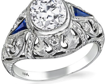 May include: A vintage 18k white gold engagement ring featuring a large, round, bezel-set diamond and two smaller blue sapphire accent stones. The ring has intricate filigree detailing.