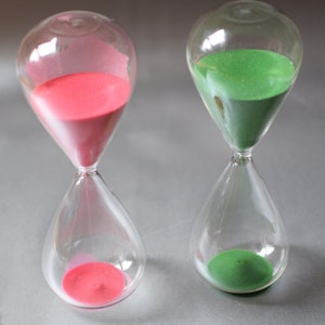 May include: Two clear glass hourglasses, one with pink sand and the other with green sand. The hourglasses have a classic teardrop shape, with the sand filling the bottom bulb. The background is a solid gray.