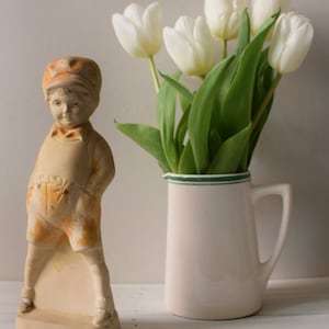 May include: A ceramic figurine of a boy wearing a cap and overalls, standing on a base. The figurine is next to a white pitcher with a green rim, filled with white tulips.