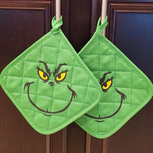 May include: Two green quilted pot holders with a cartoon face design. The face has yellow eyes, a black frown, and a wide smile. The pot holders are hanging on a wooden rod against a dark brown background.