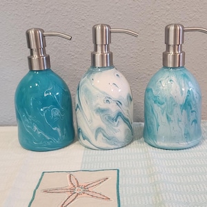 May include: Three decorative liquid soap dispensers with a marbled turquoise and white design. Each bottle has a silver pump. The dispensers are displayed on a white and blue patterned towel with a starfish design.