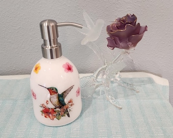 May include: A white ceramic soap dispenser with a stainless steel pump features a hummingbird and floral design. A clear glass hummingbird and a purple glass rose are also present, creating a decorative set.