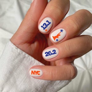 May include: White nail polish base with blue and orange decals. The thumb features the text NYC in orange. The index finger displays the number 26.2 in blue. The middle finger shows an orange Statue of Liberty with blue stars. The ring finger has 13.1 in blue with an orange star.