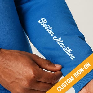 May include: A close-up of a blue sweatshirt sleeve with the words "Boston Marathon" in white script. The sleeve has a yellow and orange custom iron-on label.