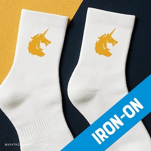 Puede incluir: Par de calcetines blancos con un diseño de cabeza de unicornio dorado. Los calcetines tienen un puño acanalado y están hechos de un material suave. Las palabras "IRON-ON" están impresas en una pancarta diagonal azul.