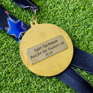 Pode incluir: Medalha dourada com uma placa retangular que diz "Tyler Tychman Run for the Shelters 5K 21:15". Pendurada em uma fita azul e preta com um padrão de estrelas. O fundo é verde.