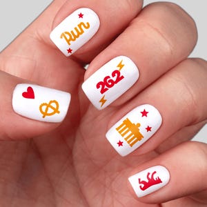 May include: White nail art with various designs. One nail features the word "Run" in orange with red stars. Another displays the number "26.2" in red. Other nails have a heart, pretzel, Brandenburg Gate, and a bear.