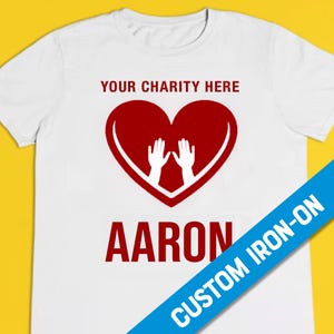 May include: White t-shirt with a red heart and two hands inside. The text "YOUR CHARITY HERE" is above the heart and the name "AARON" is below the heart. The words "CUSTOM IRON-ON" are diagonally across the bottom right corner of the shirt.