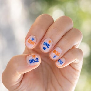 Könnte beinhalten: Nagelkunst auf hellrosa Basis mit bunten Aufklebern. Der Daumen zeigt '26.2' mit einem orangefarbenen Stern. Der Zeigefinger zeigt 'LON' mit einem blauen Herz. Der Mittelfinger hat 'NYC' mit einem blauen Herz und orangefarbenem Stern. Der Ringfinger zeigt 'BOS' mit einem blauen Blitz. Der kleine Finger zeigt 'CHI'.