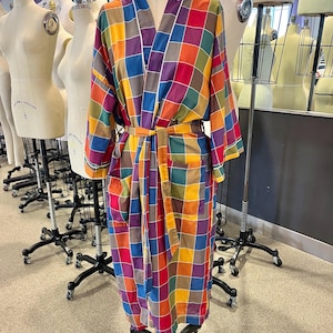May include: A colorful, checkered robe displayed on a mannequin. The robe features a vibrant patchwork design with squares of red, orange, yellow, green, blue, and purple. It has a white trim and a matching belt. The robe is displayed in a studio setting.