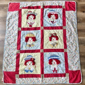 May include: A handmade quilt featuring six square panels with appliqued doll faces. Each doll has red yarn hair and wears a different hat or bow. The panels are set within red borders and surrounded by a floral patterned fabric.