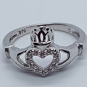 May include: A silver ring featuring a heart-shaped design with a crown on top. The heart is set with small clear stones.