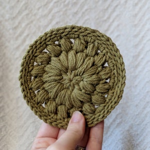 May include: A green crocheted coaster with a circular design. The coaster is made with a textured yarn and has a raised center.