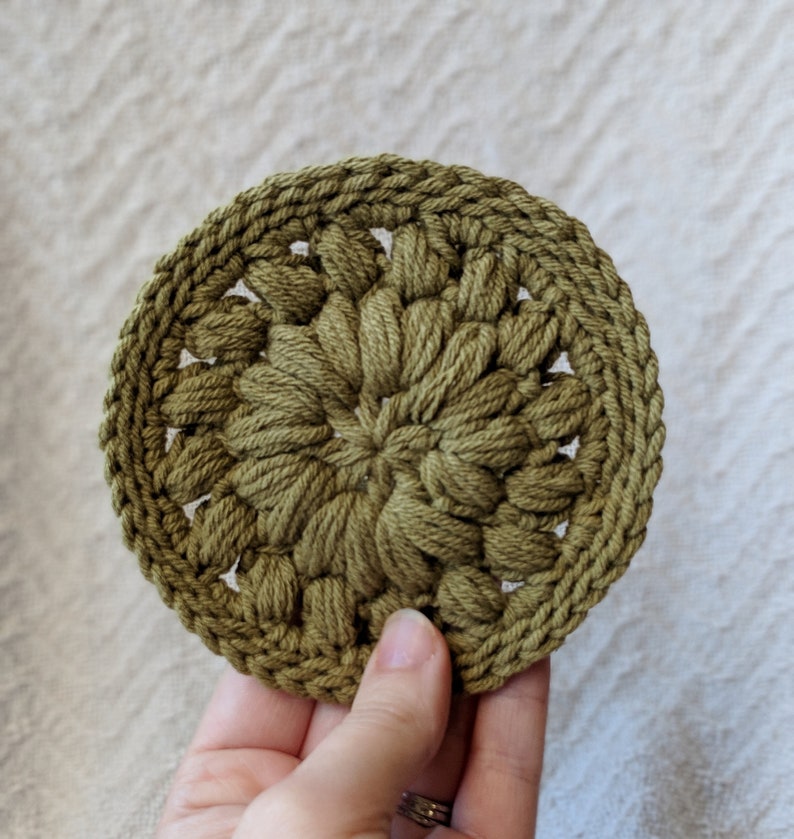 May include: A green crocheted coaster with a circular design. The coaster is made with a textured yarn and has a raised center.