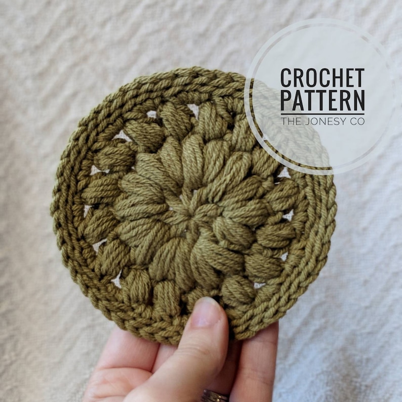 May include: A green crocheted coaster with a circular design. The coaster is made with a textured stitch that creates a raised, puffy effect. The text "CROCHET PATTERN THE JONESY CO" is visible in the image.