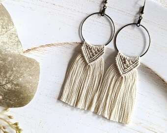 May include: A pair of cream-colored macrame earrings with a hoop design and a tassel fringe. The earrings are made with natural fibers and have a simple, bohemian style.