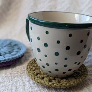 May include: A white ceramic mug with a green rim and green polka dots, sitting on a green crocheted coaster.