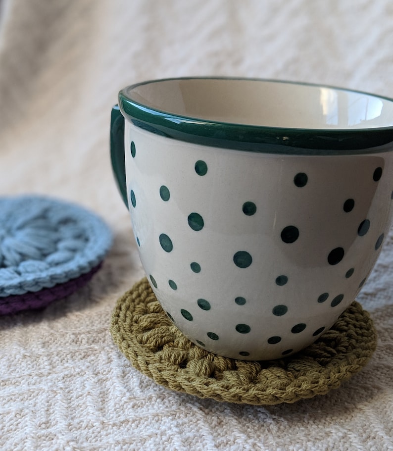 May include: A white ceramic mug with a green rim and green polka dots, sitting on a green crocheted coaster.