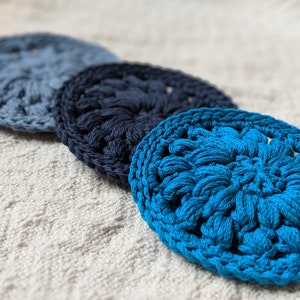 May include: Three crocheted coasters in shades of blue. The coasters are round and have a textured, floral design. The coasters are arranged in a row, with the darkest blue coaster in the middle.