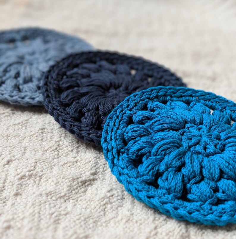 May include: Three crocheted coasters in shades of blue. The coasters are round and have a textured, floral design. The coasters are arranged in a row, with the darkest blue coaster in the middle.