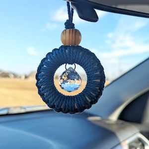 May include: A blue macrame circle with a crystal hanging from it. The circle is hanging from a wooden bead and string from the rearview mirror of a car.