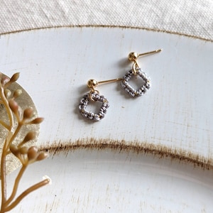 May include: A pair of silver and gold earrings with a small square shape. The earrings are made of braided wire and have a gold ball at the top.