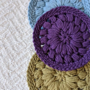 May include: Three crocheted coasters in different colors: blue, purple, and green. The coasters are round and have a floral design.