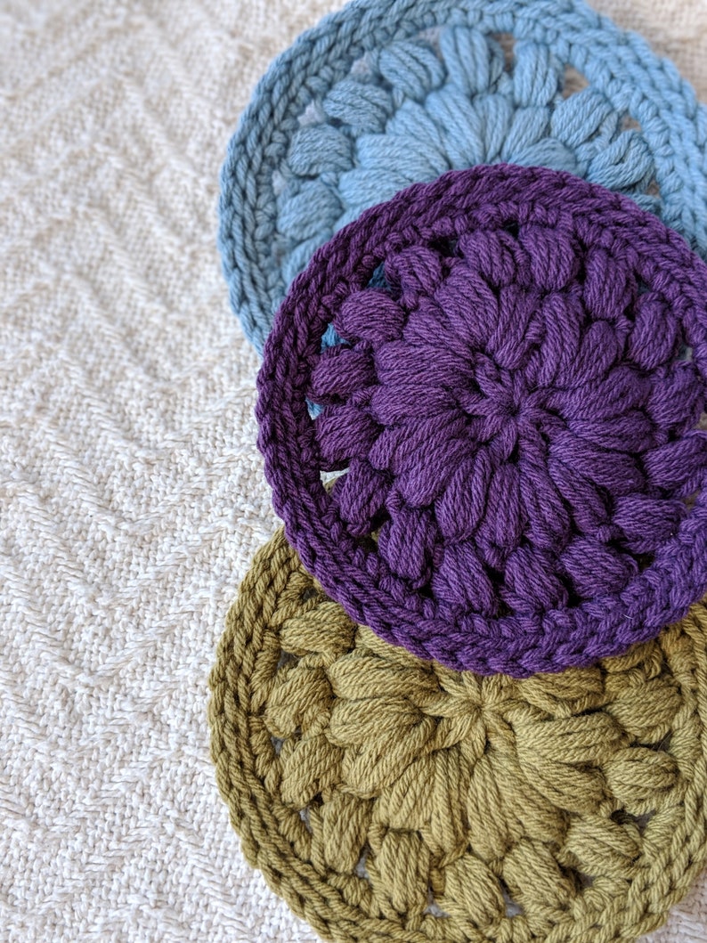 May include: Three crocheted coasters in different colors: blue, purple, and green. The coasters are round and have a floral design.