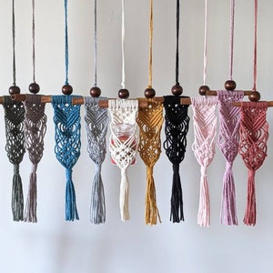 May include: Ten macrame plant hangers in various colors, including gray, blue, pink, and white, with a wooden dowel rod at the top. Each hanger has a small, clear glass jar at the bottom.