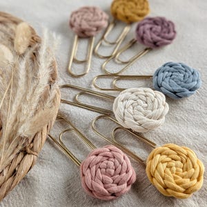 May include: A collection of decorative paper clips with a woven, floral-like design. The clips feature various colors, including pink, yellow, purple, blue, and cream, attached to gold-colored metal clips. The items are arranged on a neutral-toned fabric surface.