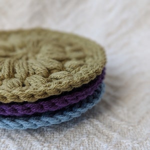 May include: A stack of three crocheted coasters in green, purple, and blue. The coasters are round and have a textured design.