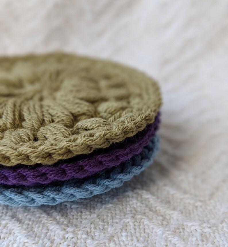 May include: A stack of three crocheted coasters in green, purple, and blue. The coasters are round and have a textured design.