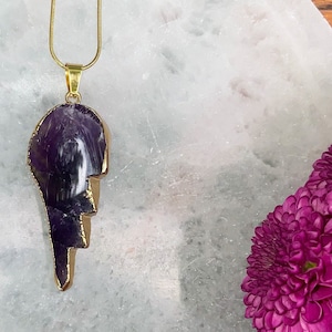 May include: A gold chain necklace with a large, purple amethyst pendant in the shape of a lightning bolt. The pendant is set against a white background.